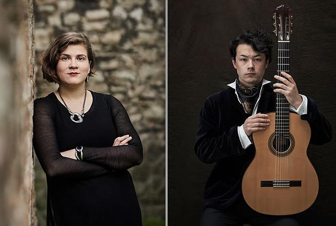 A lady wearing a black dress and silver jewellery leans against a wall, while a stoic man in black holds his guitar in front of him.