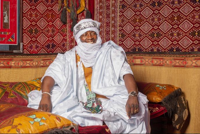 Artist in a white robe and turban seated among vibrant geometric patterned textiles and colorful cushions.