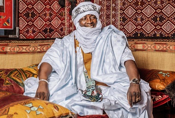 Artist in a white robe and turban seated among vibrant geometric patterned textiles and colorful cushions.