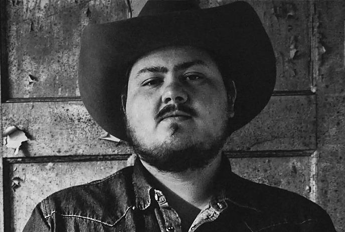 Artist in cowboy hat and denim shirt standing before rustic weathered wall in black and white.