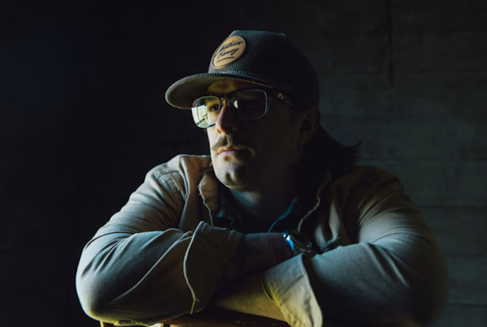 Hardy wearing a cap and glasses leans on a chair, illuminated by dramatic side lighting on a dark background.