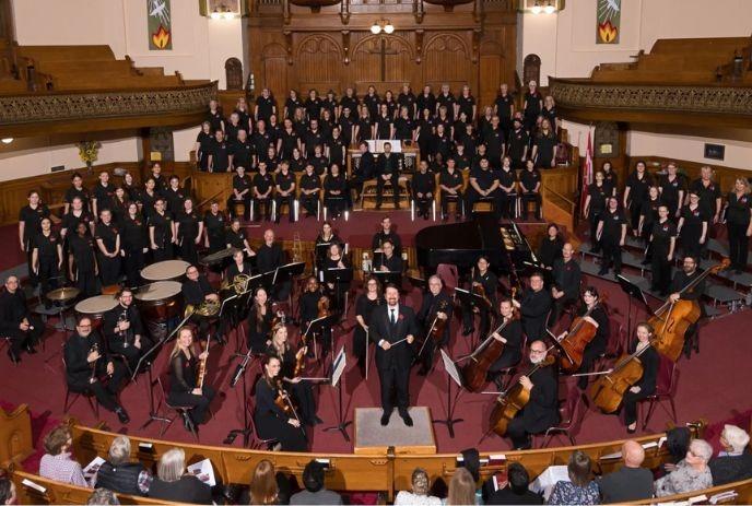 Large choir and orchestra assembled in a church sanctuary before an audience and pipe organ.