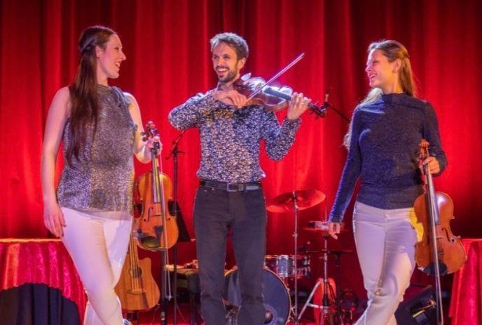 Three violinists on stage with red curtains, drum kit, and acoustic guitar behind them.