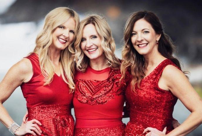 Three women artists in red sequined dresses pose together against a softly blurred backdrop.