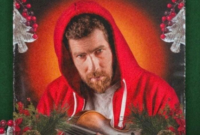Artist in a red hoodie, holding a violin amid festive holiday greenery and decorations.