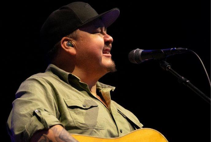 Artist sings into a microphone while playing an acoustic guitar on stage.