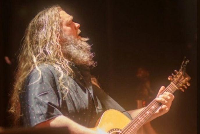 Matt Andersen with long curly hair and beard playing an acoustic guitar under warm stage lighting.
