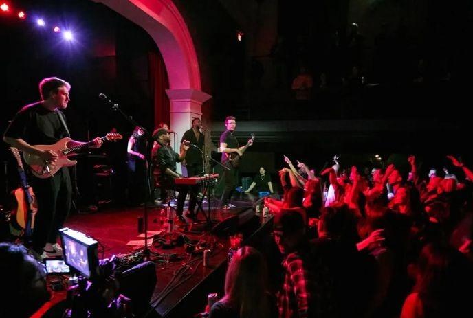 Live band performing on stage before a packed crowd, with colorful lights and energetic atmosphere.
