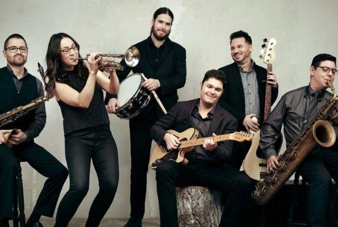 Six-piece band posing with trumpet, saxophones, drums, guitar and bass in a studio.
