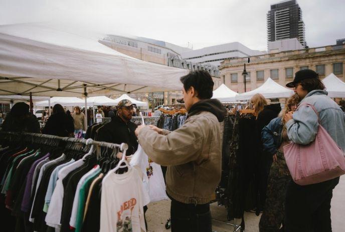 Visitors browse clothing racks beneath market tents in a busy urban square with nearby buildings.