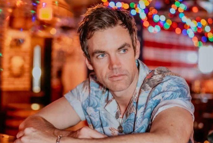 Tyler Hilton seated indoors with colorful lights in the background.