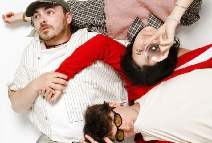 Three people lying together on a light surface, one holding a glass and one wearing sunglasses.