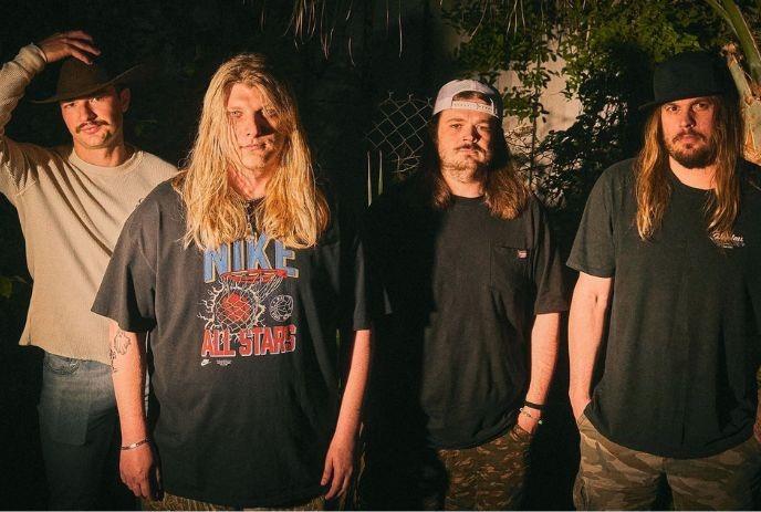 Four band members standing outdoors at night, illuminated by warm lighting.