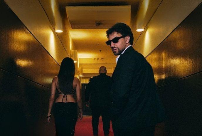 Person in dark suit and sunglasses walks a hotel hallway with two figures ahead on red carpet.