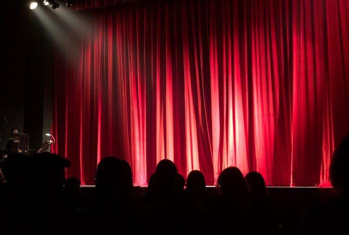 Red theatre curtain illuminated by spotlights, viewed from an audience seating area.