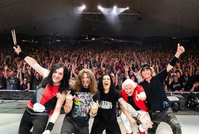 Rock band poses on stage before a packed crowd, with raised hands and bright concert lights above.