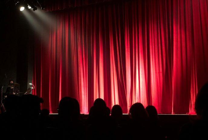 Red theater curtains illuminated by a spotlight on stage, with audience silhouettes visible in foreground.
