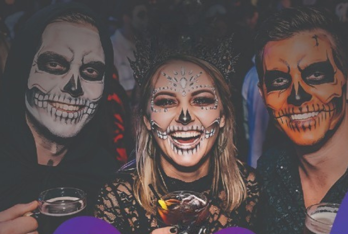 Three people with festive skeleton face paint smiling and holding drinks at a Halloween celebration.