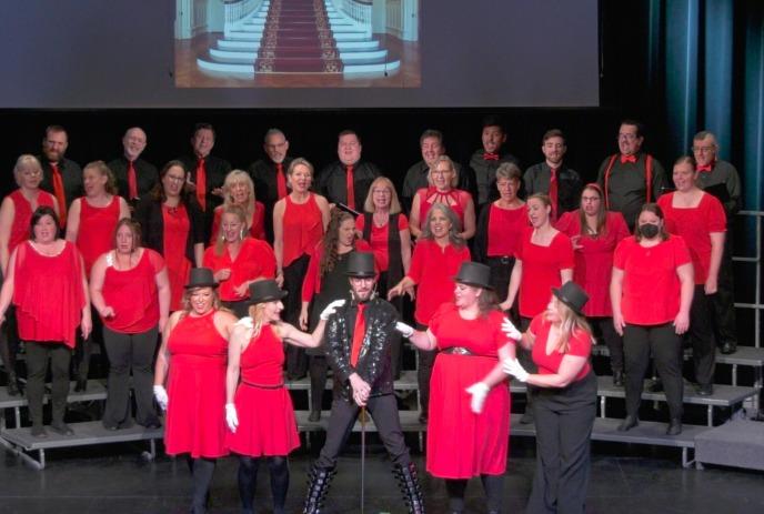 Red-clad choir performs on stage, led by a man in black leather and top hat, with a projected staircase behind.
