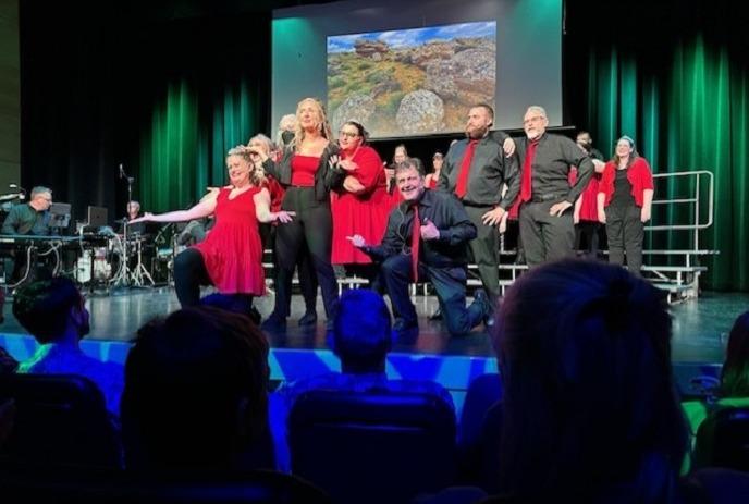 Performers in red-black costumes pose on stage; a band plays nearby, posing for the picture in front of audience.