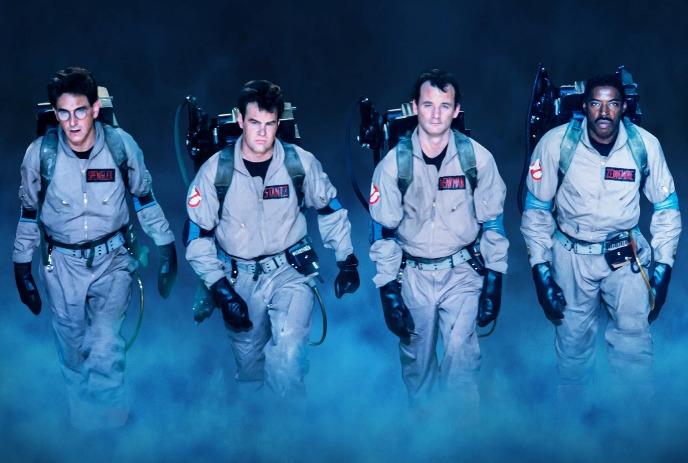 Four people wearing Ghostbuster uniforms and proton packs walk together in a foggy area.