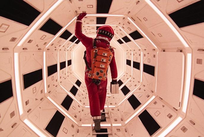Person in red spacesuit walks through bright hexagonal corridor, touching ceiling, carrying silver case.