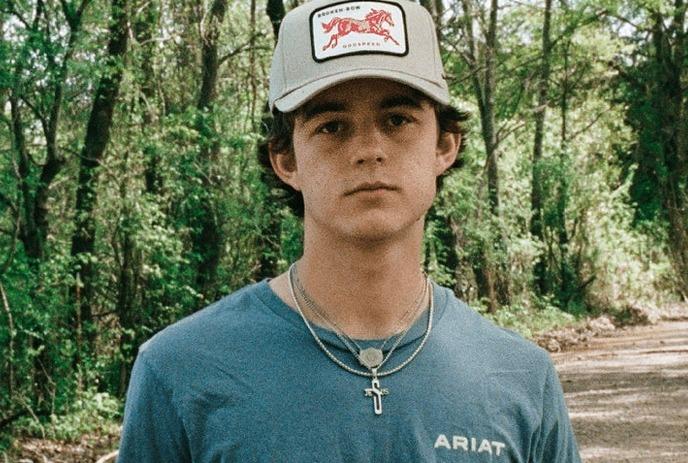 Waylon Wyatt in cap stands on dirt road amid tall green trees creating calm outdoor scene