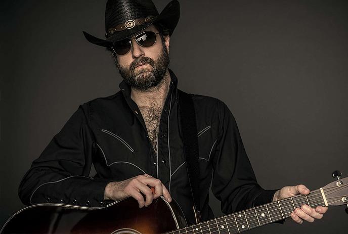 Wheeler Walker Jr. wearing a black cowboy hat, aviator shades and a beard, against a weathered wooden wall.