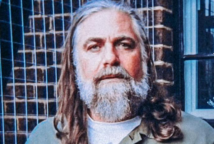 The White Buffalo with long hair wearing a green jacket stands confidently against a brick wall beside a large window.