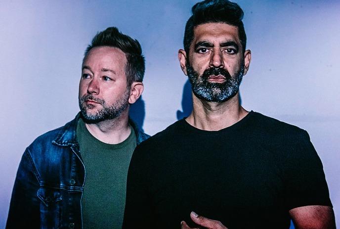 Two artists standing against blue purple backdrop wearing denim jacket and black shirt in moody lighting.