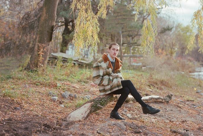 Willem James Cowan sitting on a fallen tree in autumn woods near water wearing a coat and dark clothing.