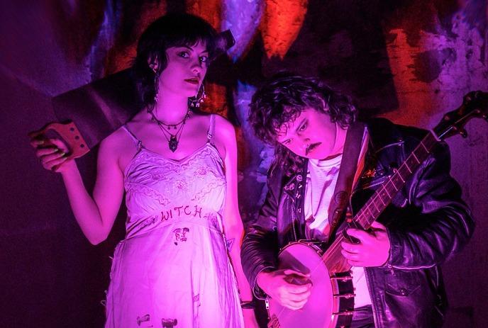 Two artists in purple lighting; one holds a banjo, and the other one is wearing a witch dress.