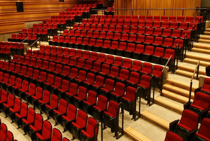 Empty auditorium with red seats and wooden paneling and central aisles leading downward.