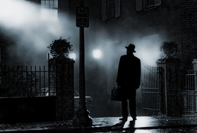Silhouetted figure in hat and coat stands on foggy sidewalk at night, gazing at brightly lit house behind iron fence.