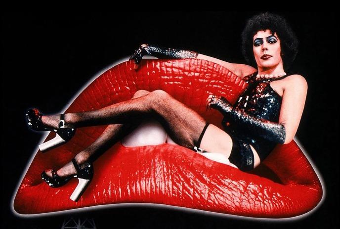 Person reclines on giant red lips, wearing sequined corset, and heels in theatrical pose.