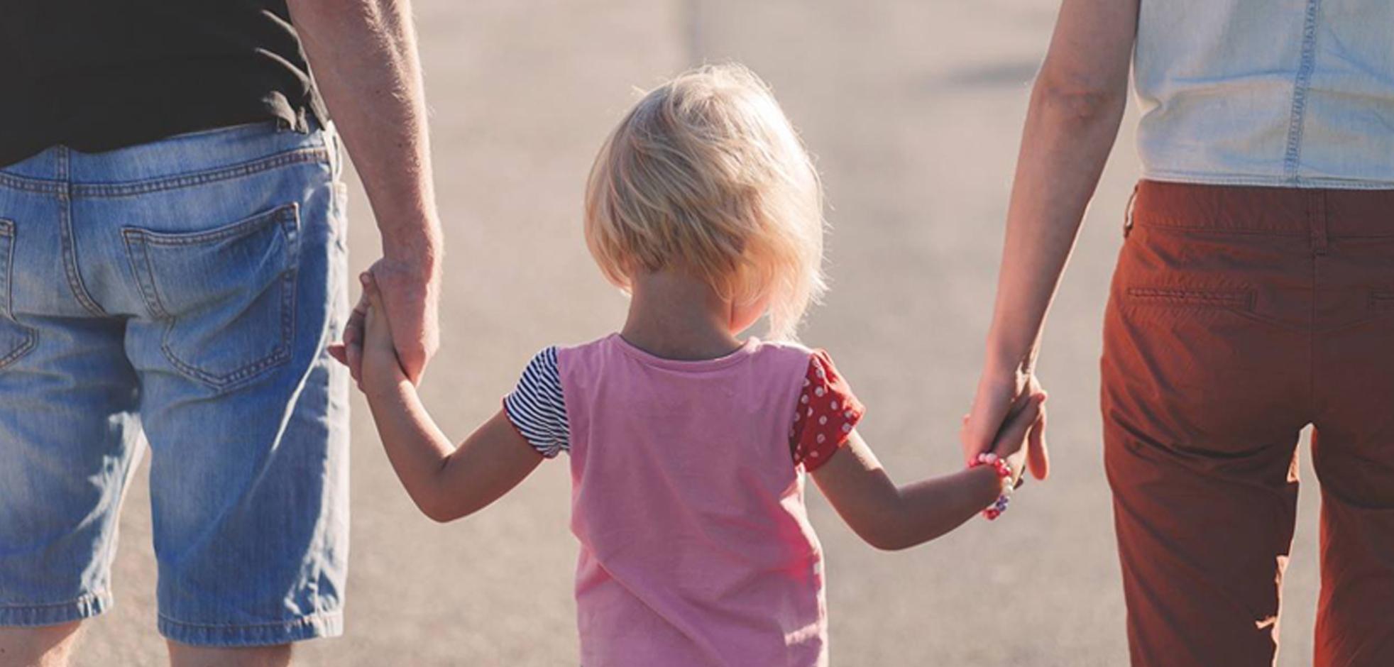 Young child holding both their left and right hand with two adults walking on a sunny day