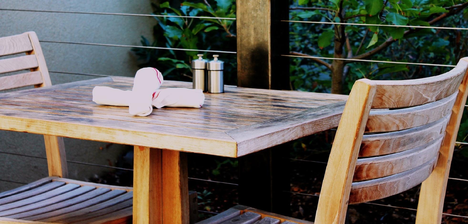 Wooden table and chairs set with cutlery and salt and pepper shakers on a patio with bushes
