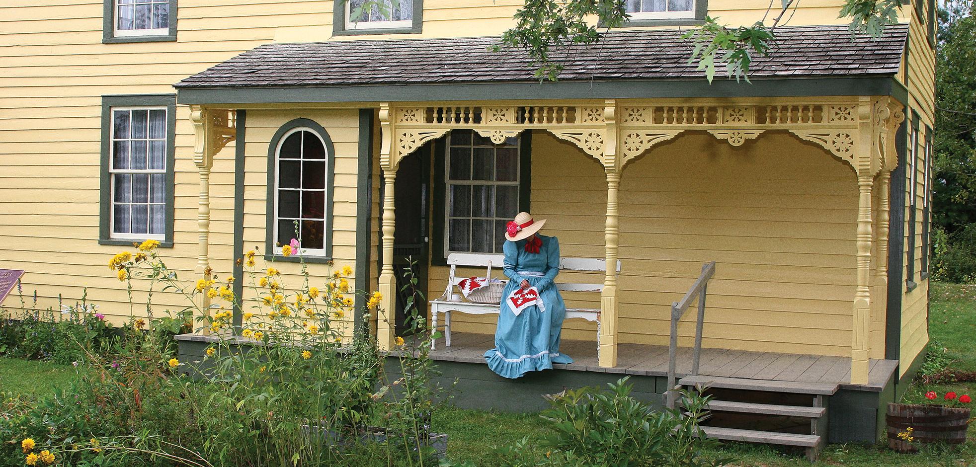 Lady in blue dress sitting on the porch of yellow house.