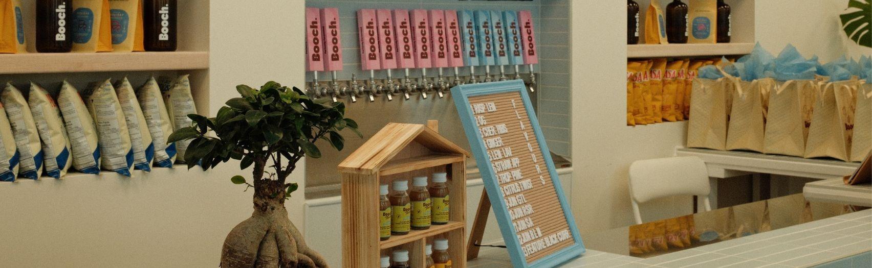 Interior of a kombucha bar with a tiled counter, bonsai, menu board, and a wall of numbered Booch taps .