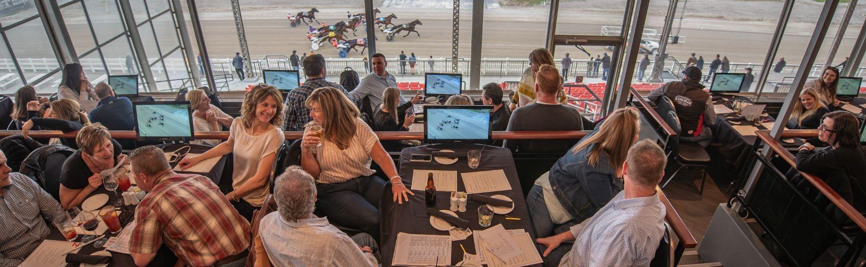 Guests watch a live horse race from an indoor lounge, dining and tracking bets as horses sprint outside.