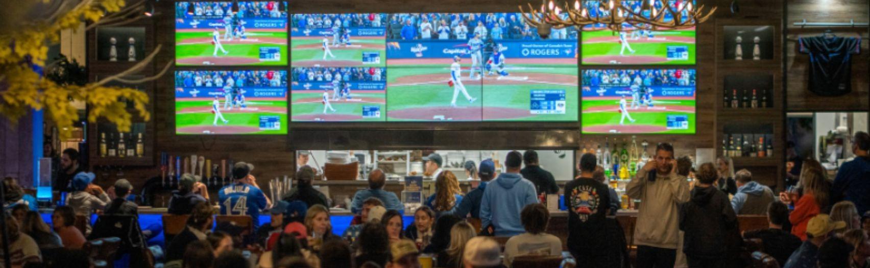 Crowded bar of fans watching a baseball game on multiple screens, with people eating, drinking, and cheering