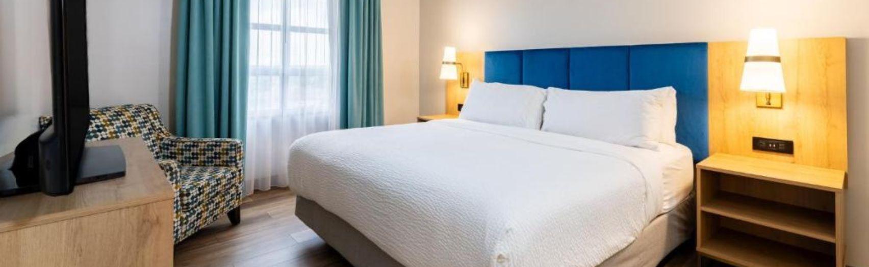 Modern room with white bed, blue headboard, patterned chair, wood furniture, and teal window drapes.