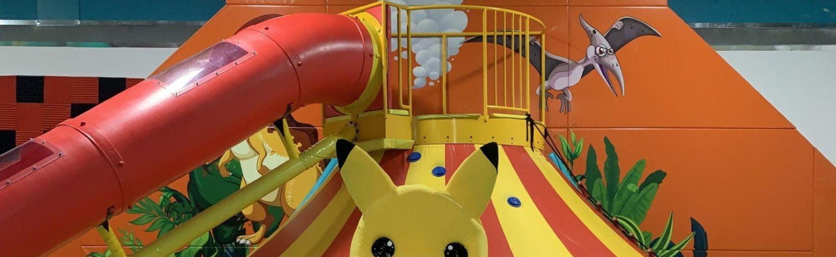 Bright indoor play area with large yellow character by a volcano slide and bold cartoon decor.