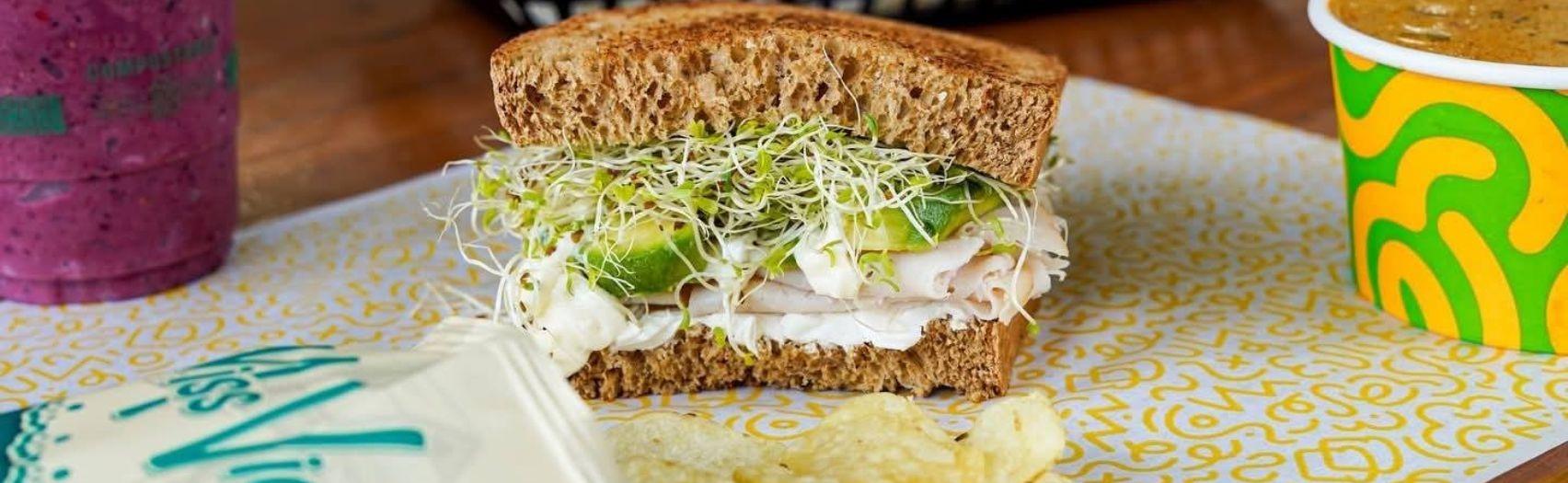 Turkey avocado sprout sandwich with chips, purple smoothie, and soup on a patterned tray.