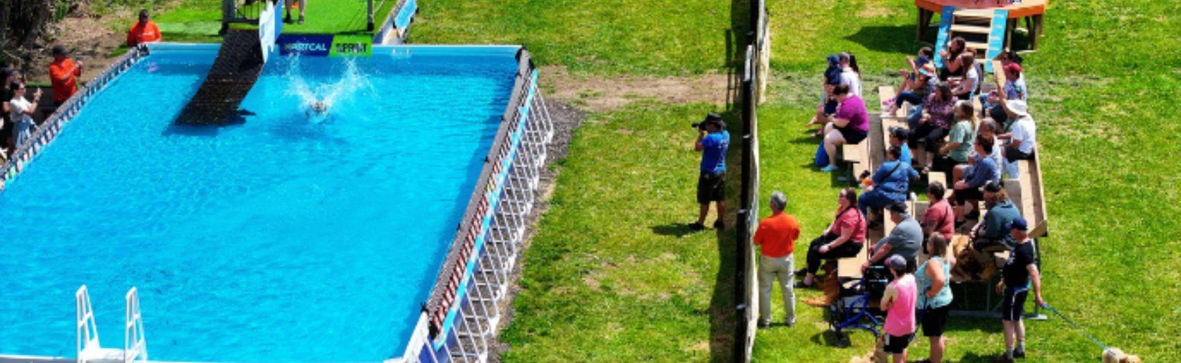Dog leaps into long pool as spectators watch from bleachers at an outdoor dock diving event.