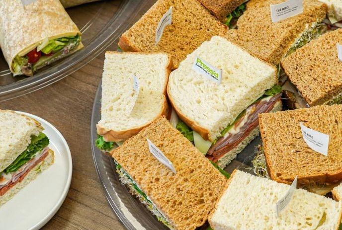 Trays of assorted labeled sandwiches and a veggie wrap arranged neatly on a wooden catering display.