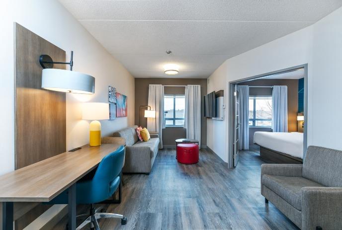 Modern suite with sofa desk chair lamp art and bed visible through wide doorway with bright window light.