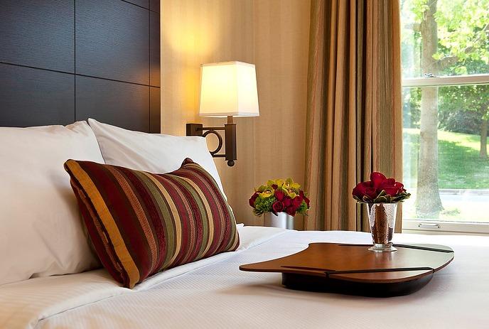 A hotel room with a neatly made bed, wooden headboard, desk, armchair, and TV on a patterned carpet.