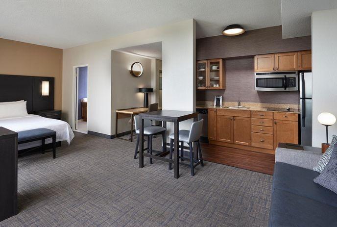 A hotel suite featuring a sleeping area with a bed, a small dining space with a table and chairs, and a kitchenette.