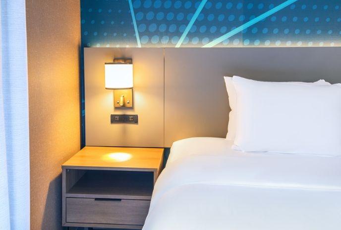 Modern hotel room bedside with white linens, wood nightstand, warm lamp, outlets, and blue patterned wall.